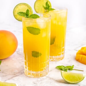 2 mango mocktails in tall glasses garnished with al lime wheel and sprig of mint on a grey and white marble background
