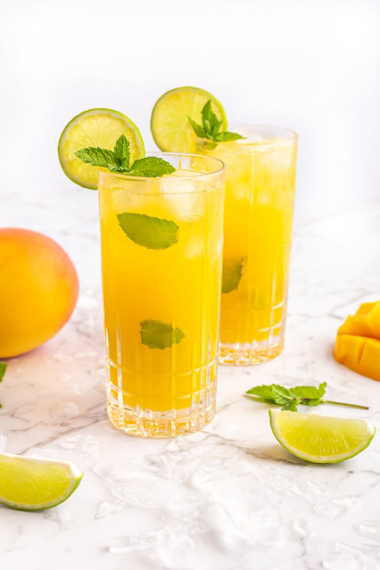 2 mango mocktails in tall glasses garnished with al lime wheel and sprig of mint on a grey and white marble background