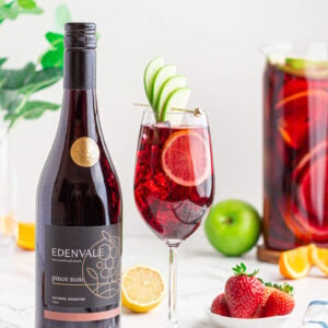 non alcoholic sangria mocktail in a wine glass garnished with an apple fan and lemon round with a jug of Sangria and a bottle of Edenvale pinot noir