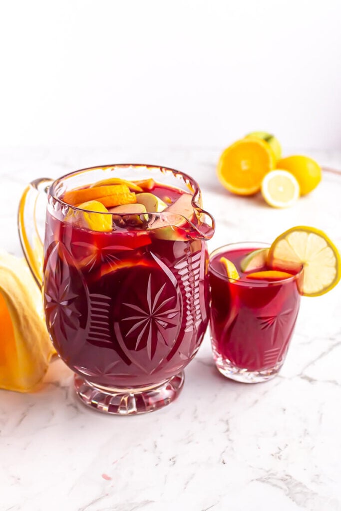 crystal jug of non alcoholic sangria garnished with orange, apple and lemon with a small glass beside it