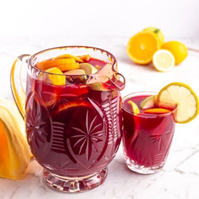 crystal jug of non alcoholic sangria garnished with orange, apple and lemon with a small glass beside it
