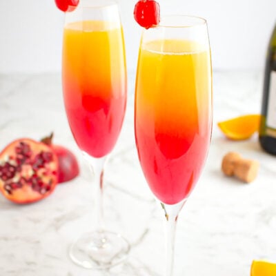 sunrise mocktail in champagne glass garnished with a cherry with pomegranate and orange in the background