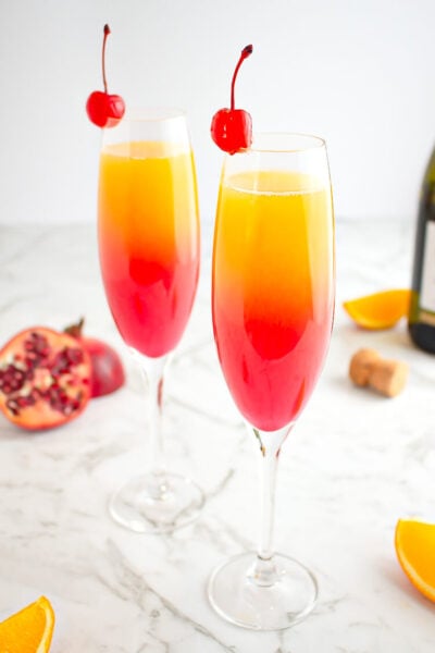 sunrise mocktail in champagne glass garnished with a cherry with pomegranate and orange in the background