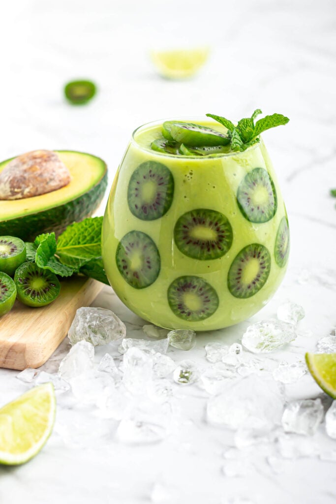 avocado mock ail garnished with kiwi berries in a round glass with avocado, kiwi berries and mint in the background