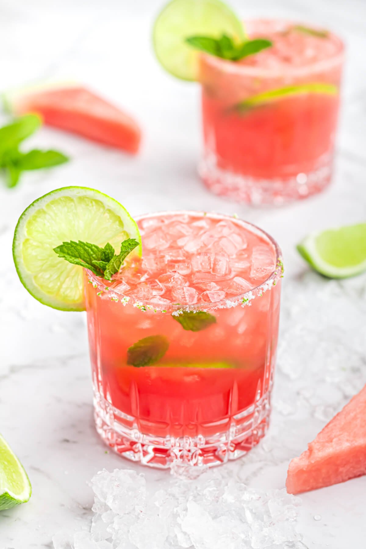 2 watermelon mocktails in short glasses garnished with mint and lime with crushed ice and lime wedges scattered in the background.