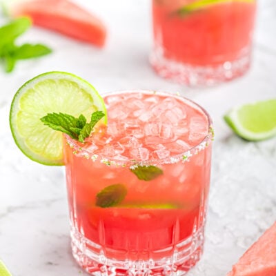 2 watermelon mocktails in short glasses garnished with mint and lime with crushed ice and lime wedges scattered in the background