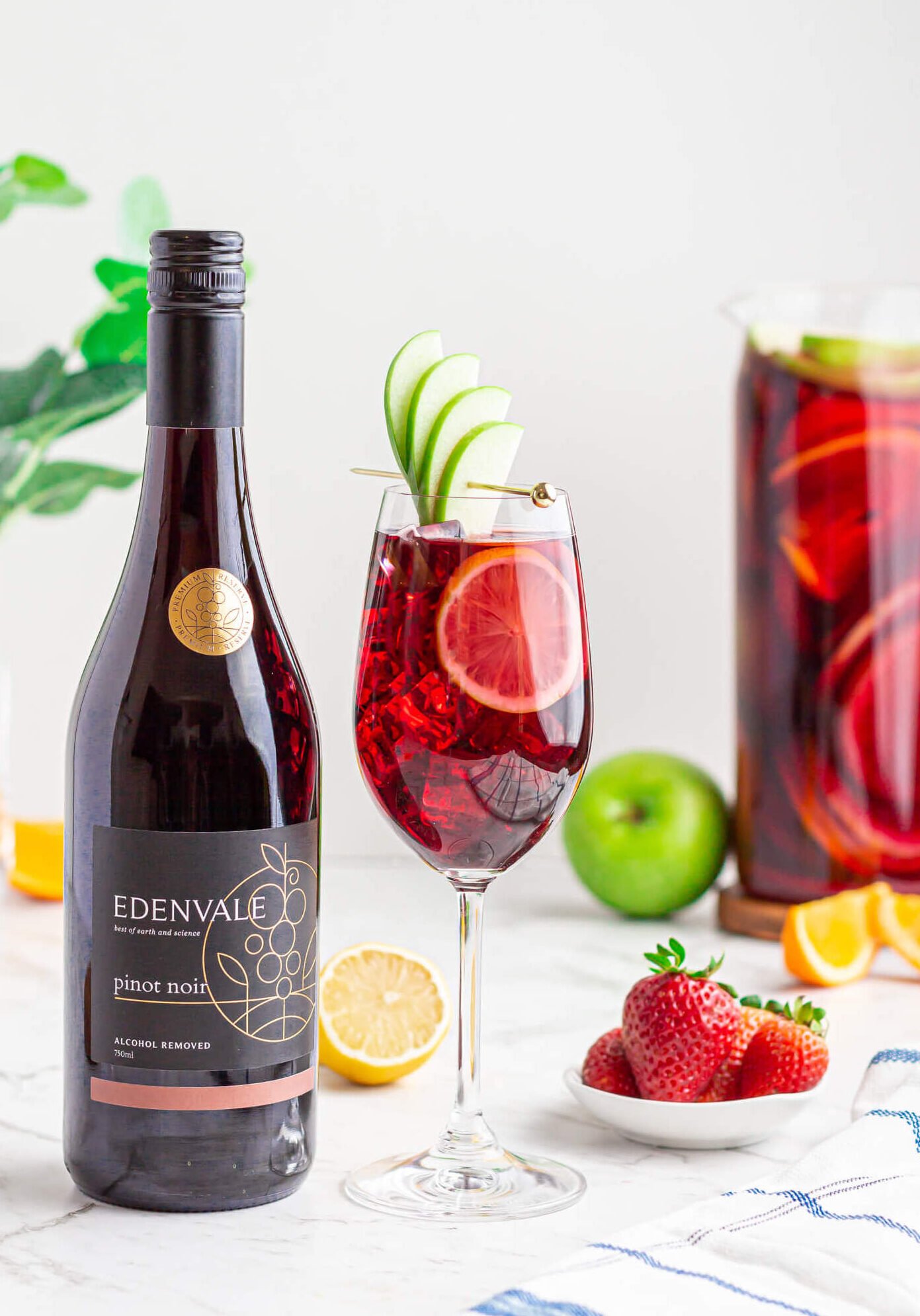 non alcoholic sangria mocktail in a wine glass garnished with an apple fan and lemon round with a jug of Sangria and a bottle of Edenvale pinot noir