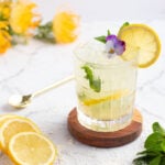 yellow mocktail in a short glass with ice on a wooden coaster garnished with mint and lemon
