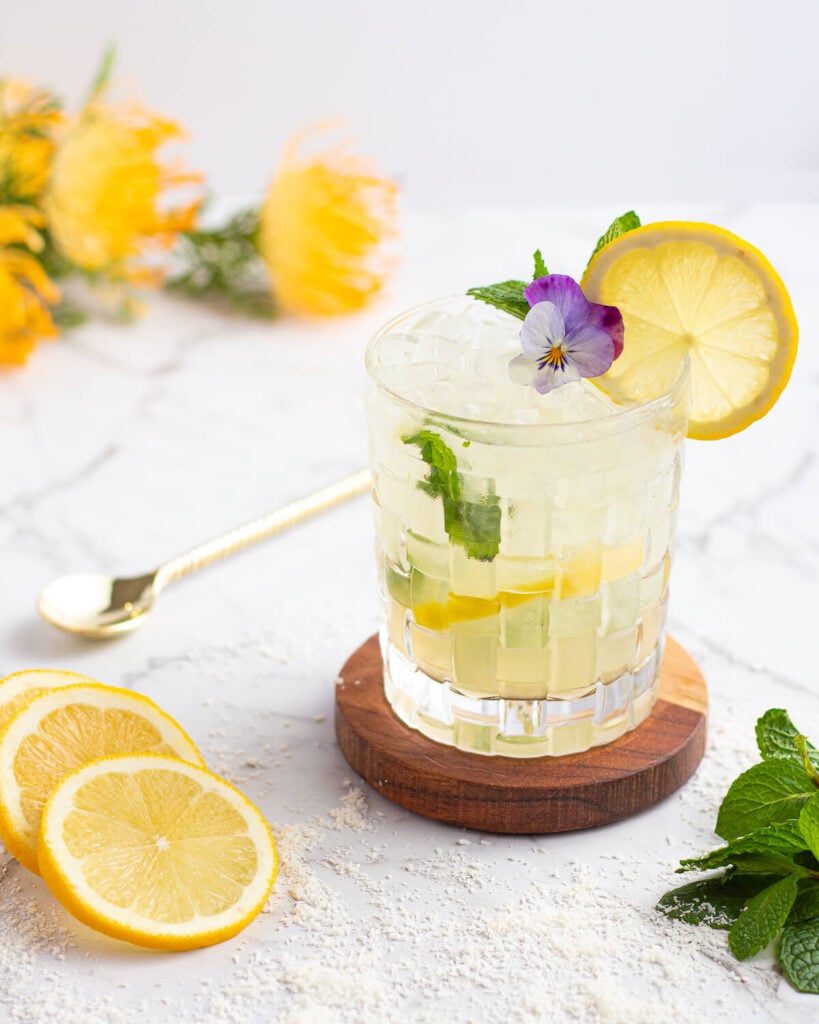 yellow mocktail in a short glass with ice on a wooden coaster garnished with mint and lemon