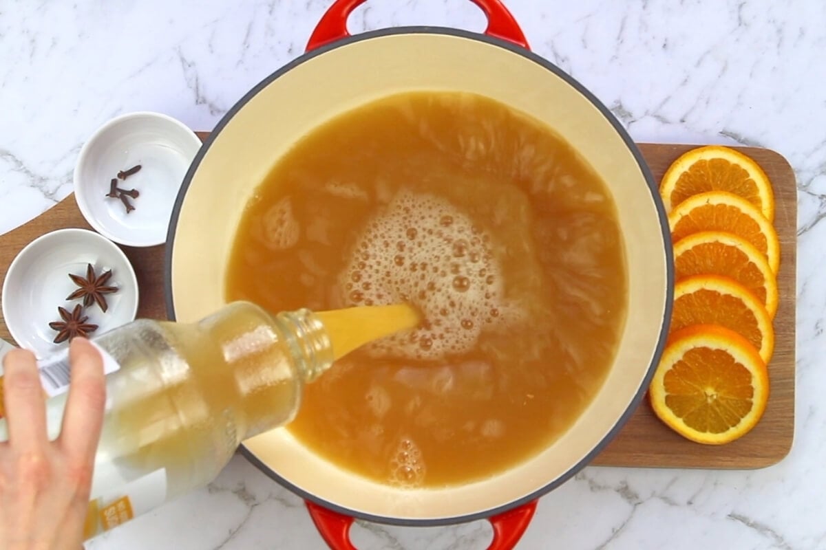 A hand pours a light-colored liquid from a bottle into a pot of mulled apple cider. Sliced oranges, star anise, and cloves are arranged on a wooden board nearby, all set on a marble countertop.