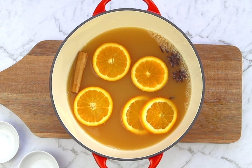 A pot filled with mulled apple cider, garnished with orange slices, a cinnamon stick, and star anise, sits on a wooden cutting board atop a marble surface. Small white bowls are placed nearby.