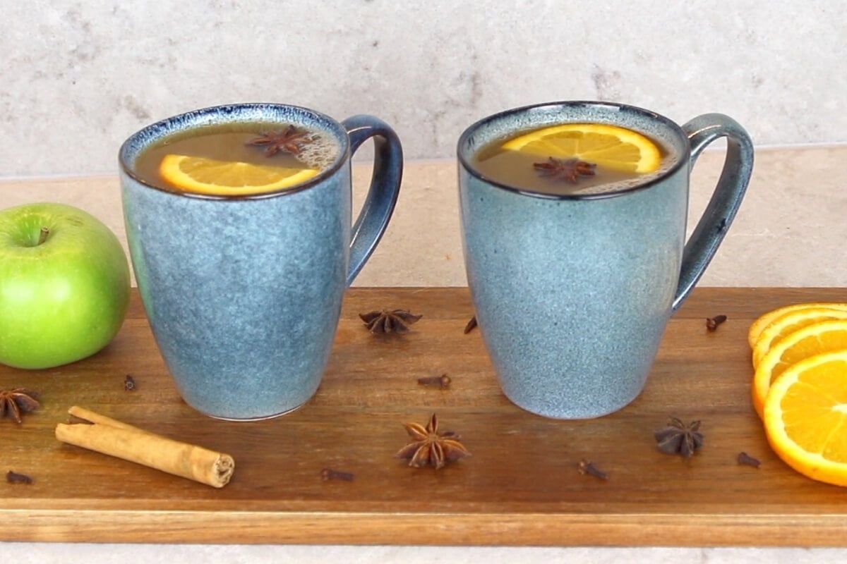 Two blue mugs filled with mulled apple cider, garnished with orange slices and star anise, sit on a wooden board. Nearby are a green apple, cinnamon stick, cloves, star anise, and orange slices.
