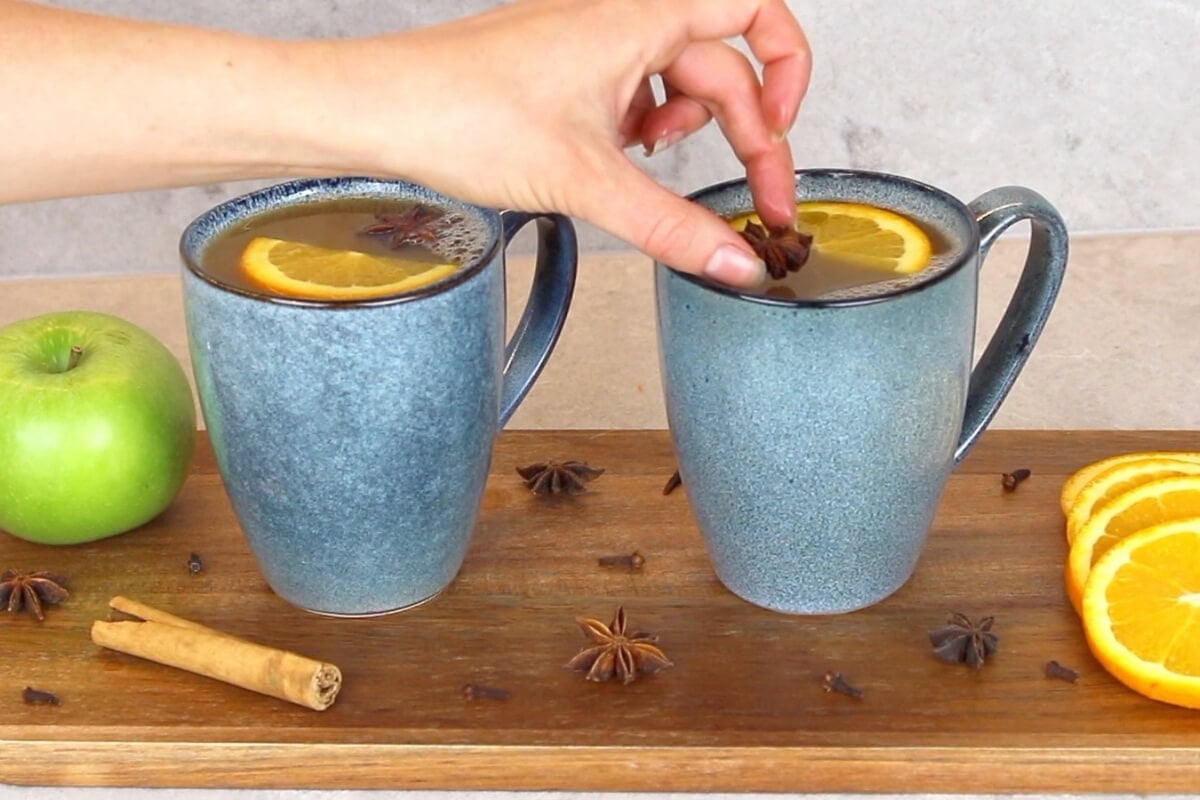 A hand places a star anise into a blue mug of mulled apple cider garnished with an orange slice. Another similar mug, apple, cinnamon stick, cloves, and more orange slices rest on a wooden board.