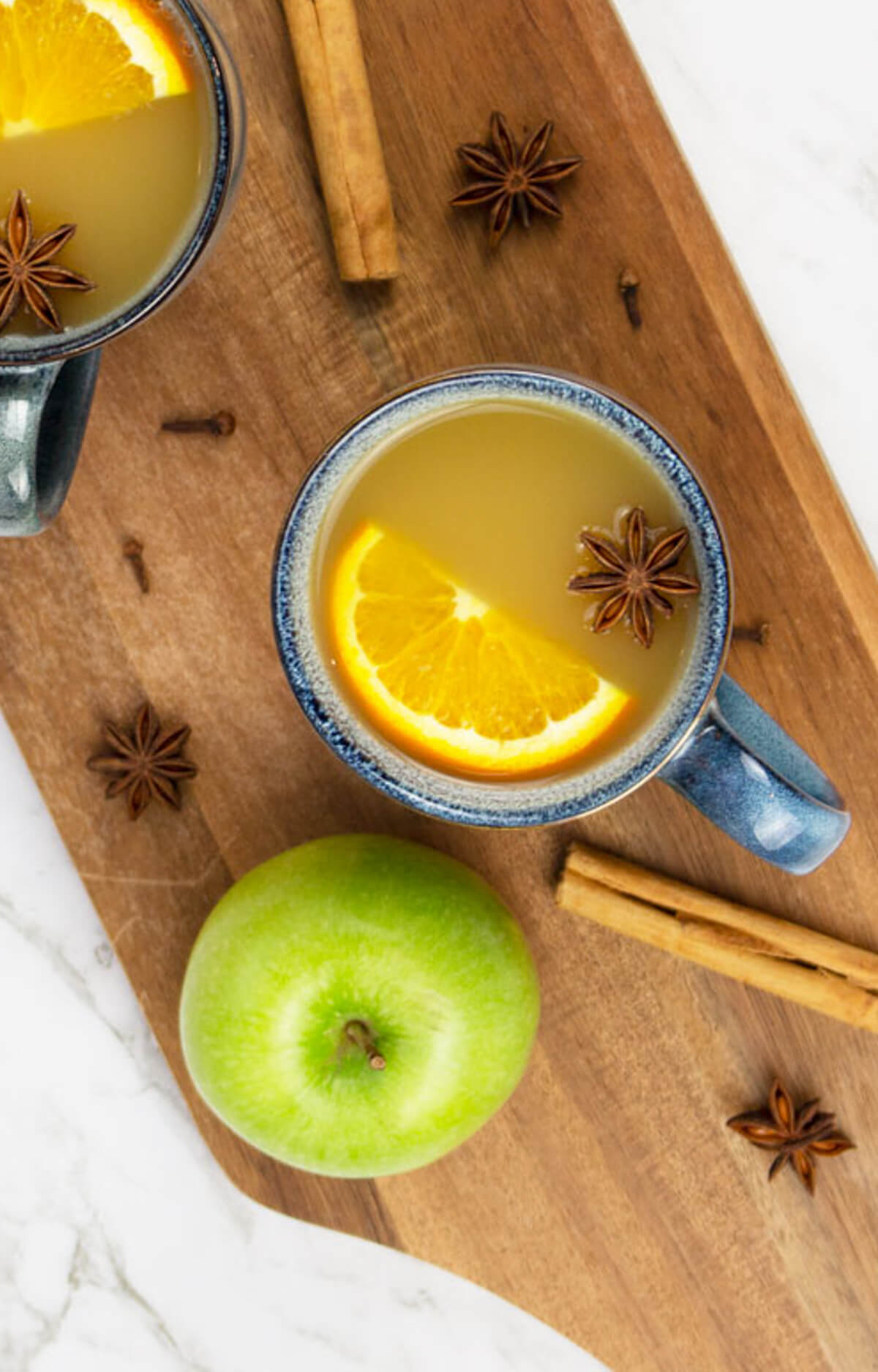 birds eye view of mulled apple cider in blue mugs on a wooden board with a green apple cinnamon sticks and star anise scattered nearby.