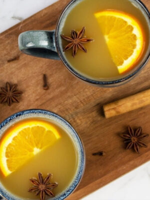 Two mugs of mulled apple cider topped with orange slices and star anise sit on a wooden board, surrounded by cinnamon sticks, cloves, an apple, and orange slices.