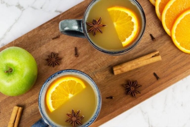 Two mugs of mulled apple cider topped with orange slices and star anise sit on a wooden board, surrounded by cinnamon sticks, cloves, an apple, and orange slices.
