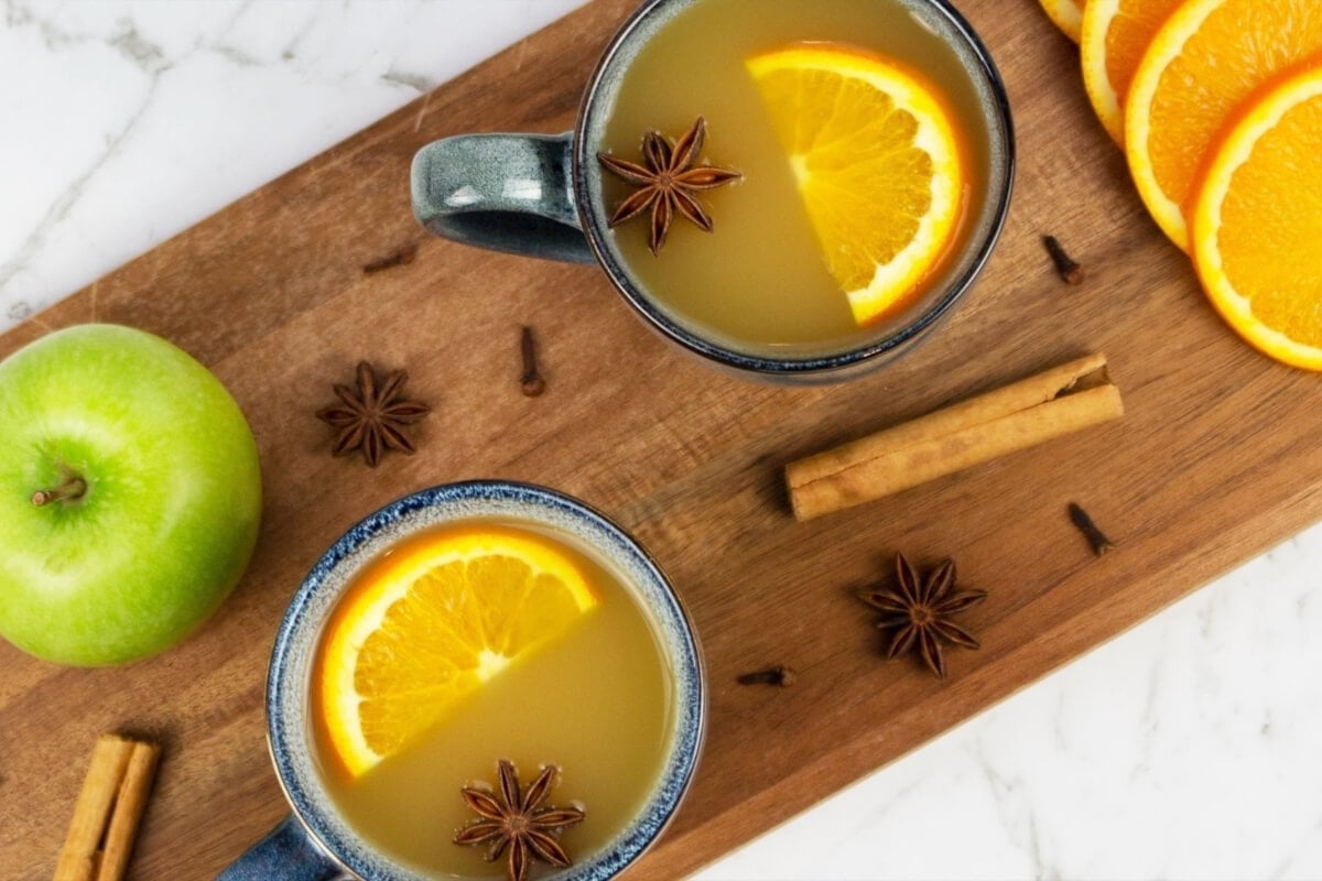 Two mugs of mulled apple cider topped with orange slices and star anise sit on a wooden board, surrounded by cinnamon sticks, cloves, an apple, and orange slices.