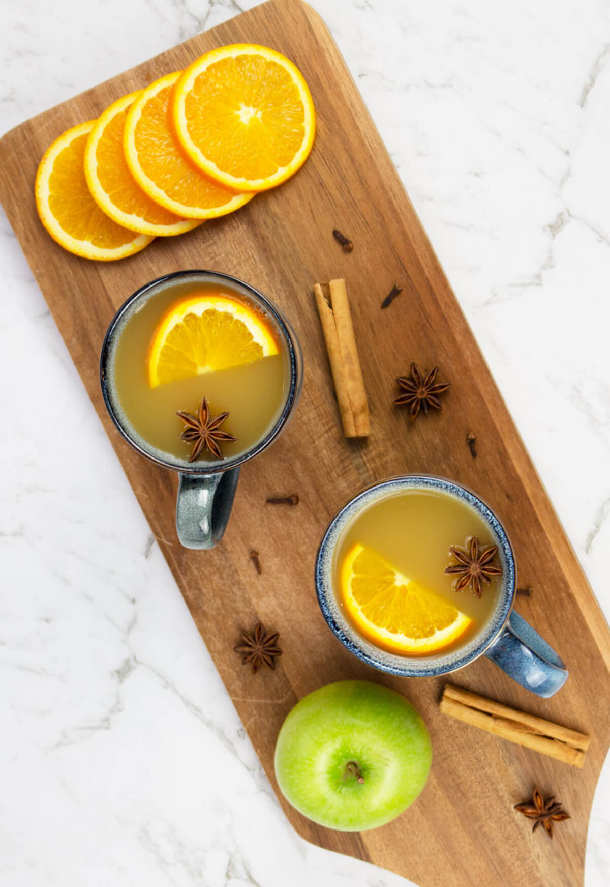 wooden board with two blue mugs of mulled apple cider garnish with an orange slice and star anise and a green apple orange slices ad spices scattered nearby.