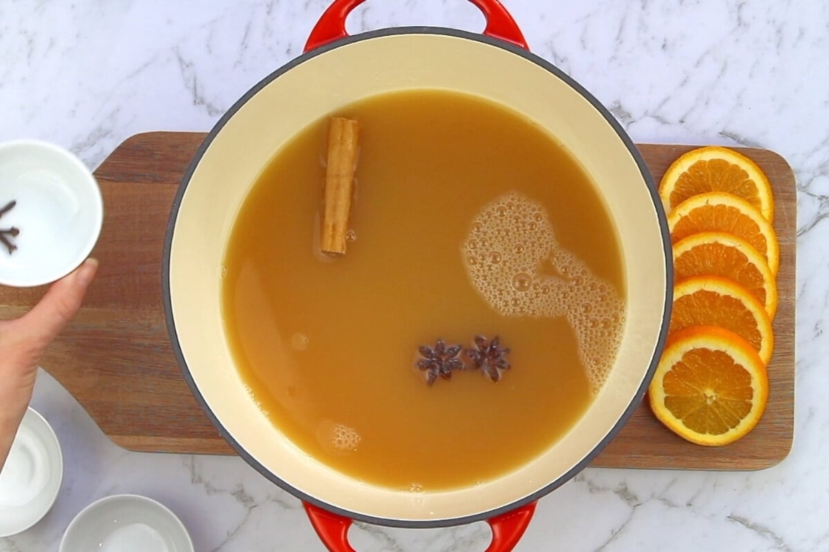 A large pot of mulled apple cider simmers with a cinnamon stick and star anise. Slices of orange rest on a cutting board, while a hand holds a small bowl of cloves over the fragrant cider.