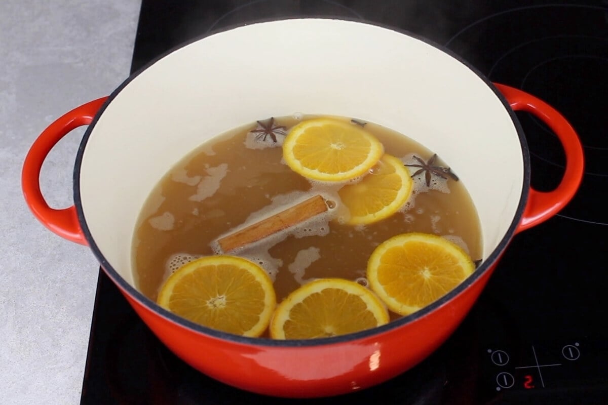 A red pot on a stovetop contains simmering mulled apple cider, with orange slices, star anise, and a cinnamon stick floating on top.