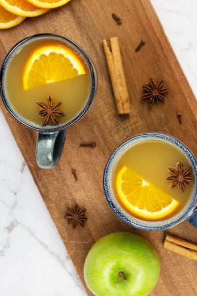 two mugs of mulled apple cider sitting on a brown wooden board with scattered spices orange slices and a green apple nearby.
