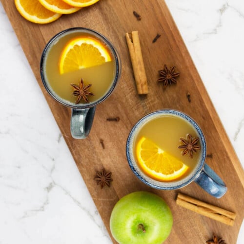 two mugs of mulled apple cider sitting on a brown wooden board with scattered spices orange slices and a green apple nearby.