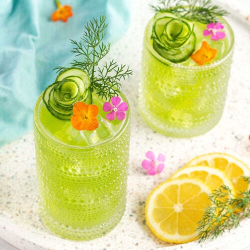 green drink in a tall glass garnished with a cucumber peel in the shape of a flower and dill