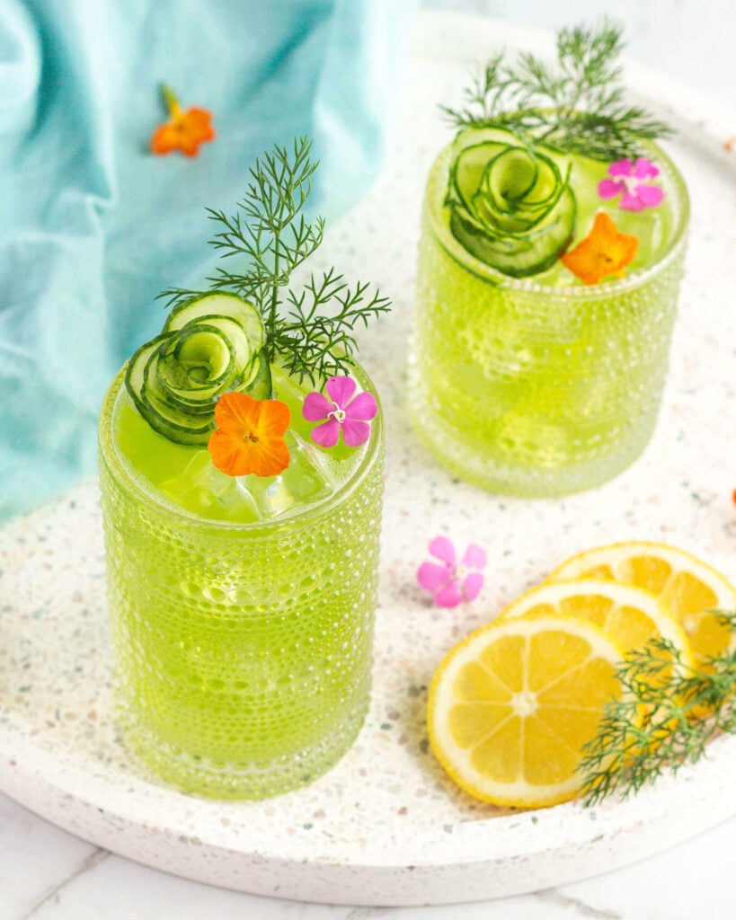 green drink in a tall glass garnished with a cucumber peel in the shape of a flower and dill
