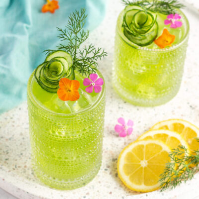 green drink in a tall glass garnished with orange and pink flowers, dill and a cucumber shaped like a flower