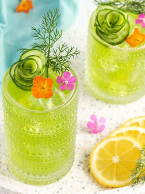 green drink in a tall glass garnished with orange and pink flowers, dill and a cucumber shaped like a flower