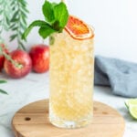 tall glass with light orange liquid and small ice blocks garnished with a citrus wheel and mint leaves and a bamboo straw