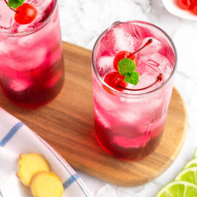 red drink in a tall glass garnished with cherries and mint served on a dark wooden board