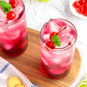red drink in a tall glass garnished with cherries and mint served on a dark wooden board