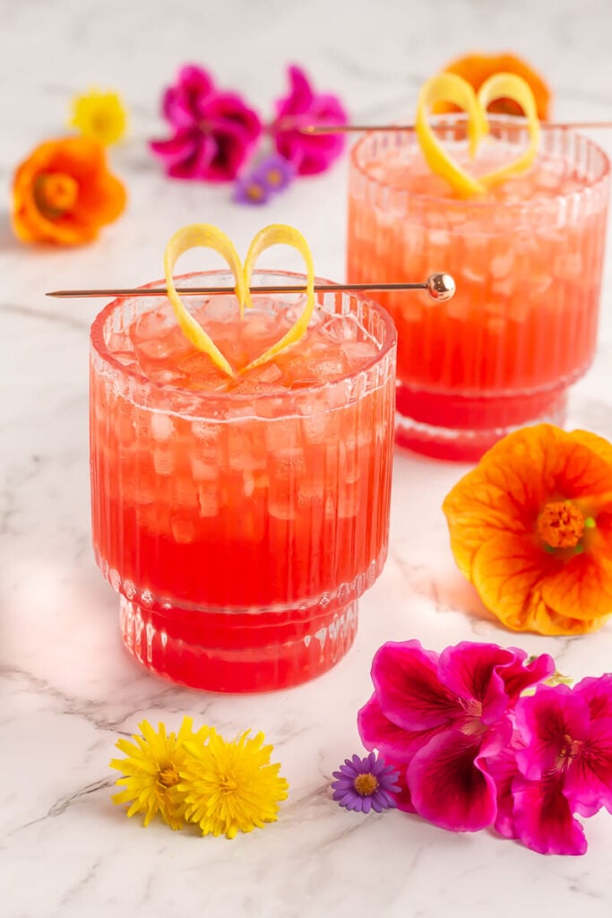 two short glasses of pink liquid surrounded by pink and orange flowers garnish with lemon peel cut into a heart shape.