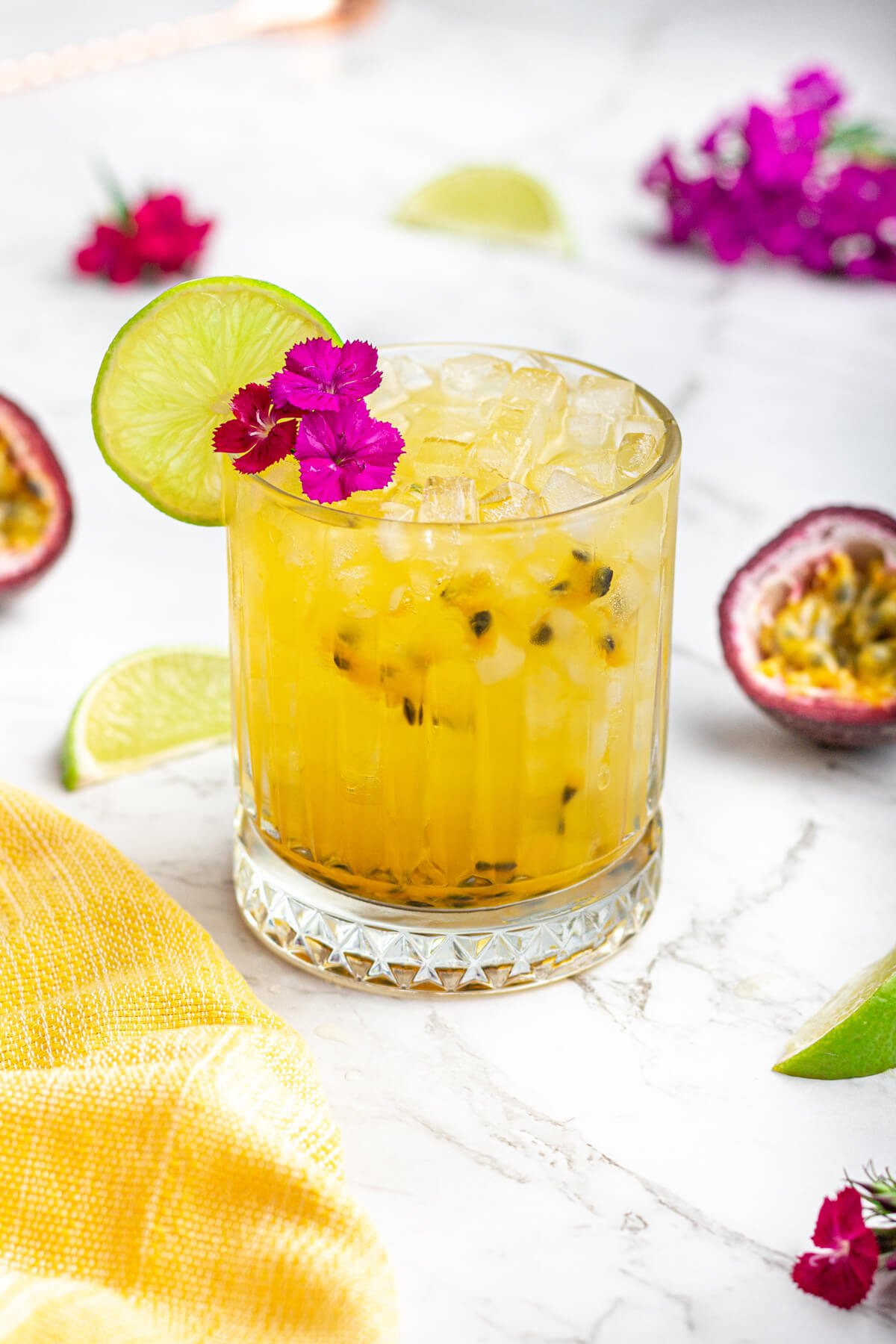 yellow drink in short glass with fresh passionfruit seeds garnished with a pink flower and lime wheel.