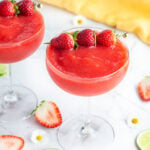 2 red strawberry daiquiri mocktails garnished with 3 small strawberries with strawberry, daises and lime scattered in the background.