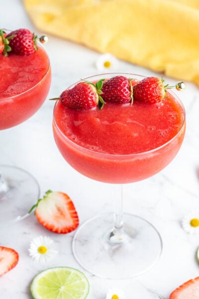 2 red strawberry daiquiri mocktails garnished with 3 small strawberries with strawberry, daises and lime scattered in the background.