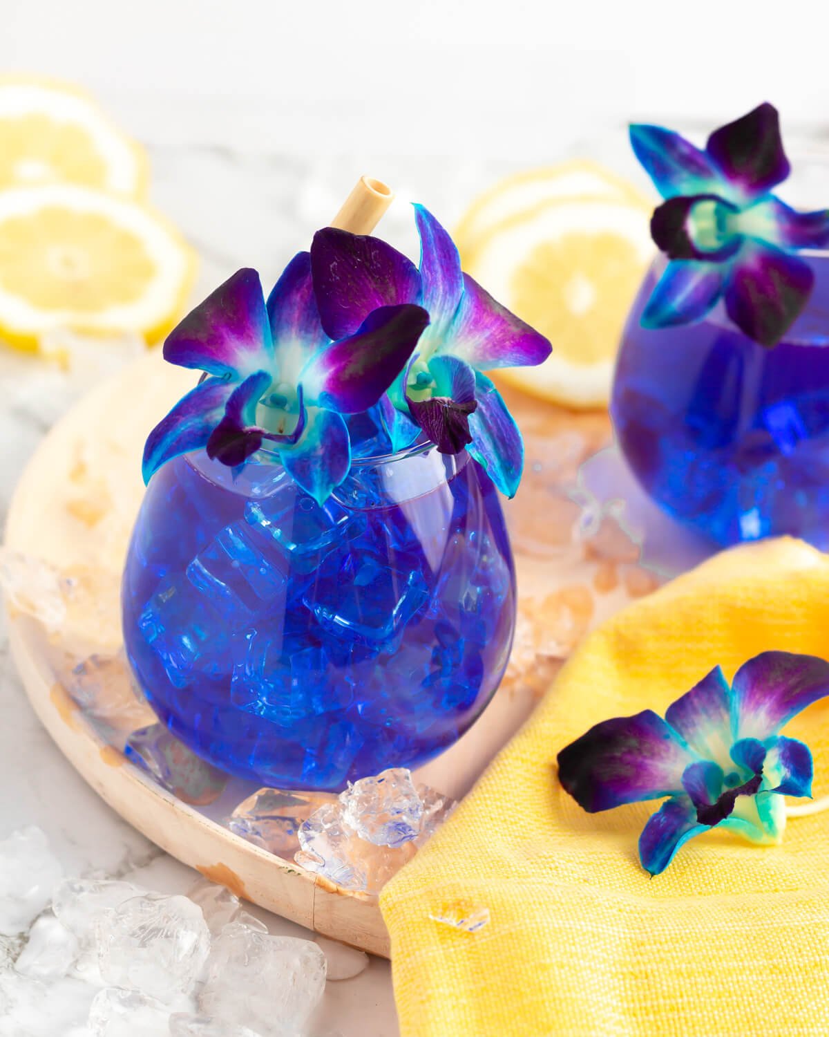 two vibrant blue lagoon mocktails garnished with blue and purple flowers on a wooden tray with ice and lemon scattered nearby.