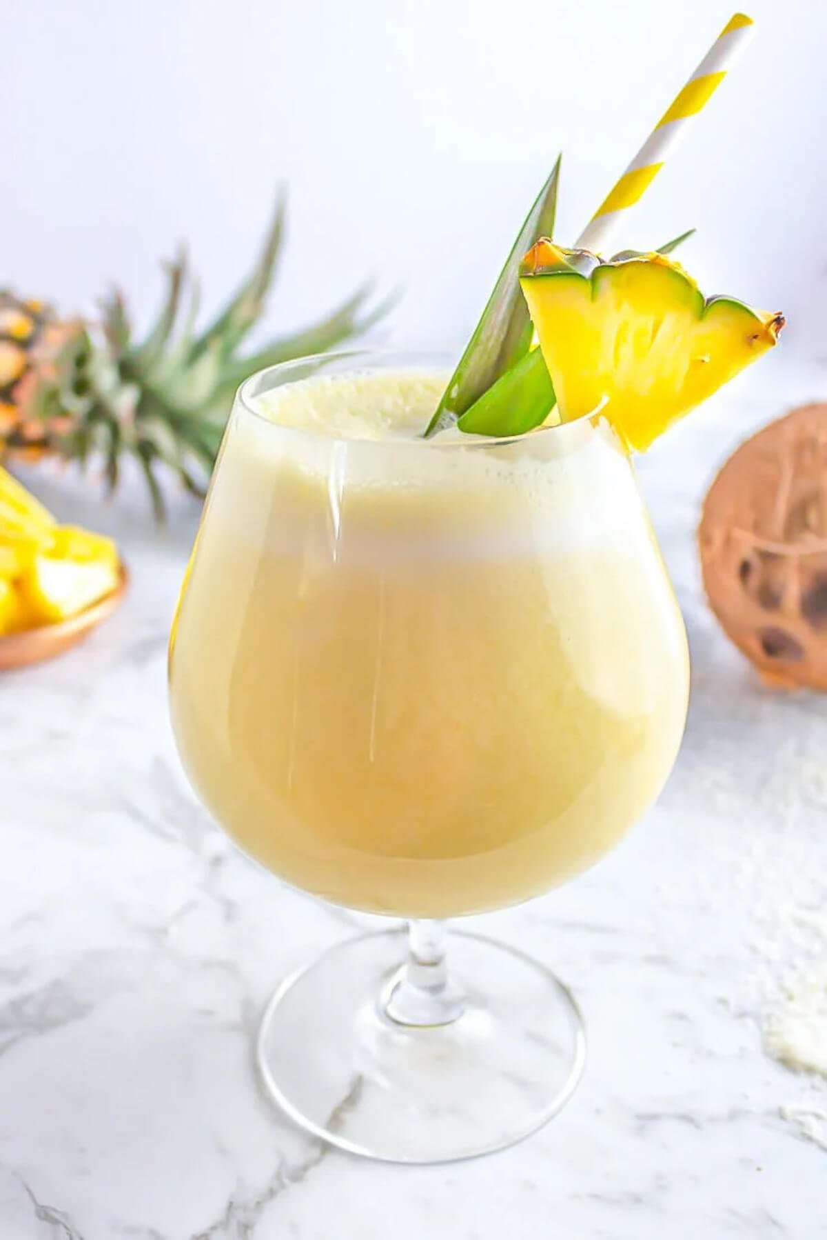 yellow virgin piña colada in a round glass garnished with fresh pineapple and fronds with a coconut and pineapple in the background.