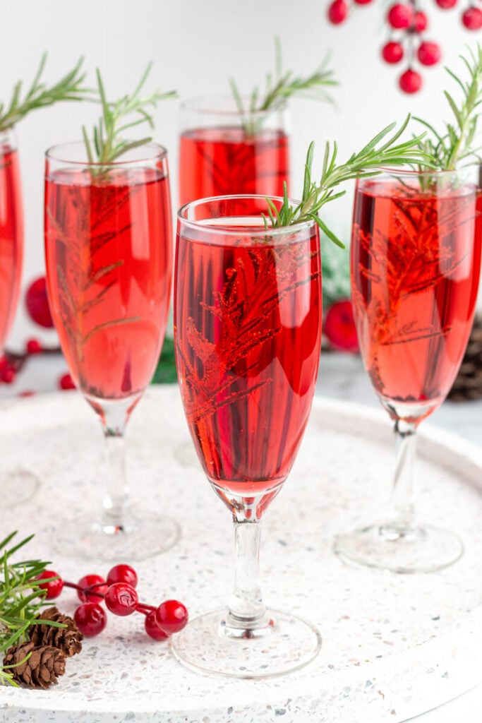 5 red mimosa drinks in champagne flutes garnished with a sprig of rosemary