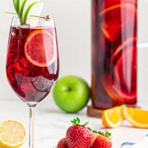 wine glass filled with non-alcoholic Sangria garnished with a green apple fan with a jug of Sangria in the background.