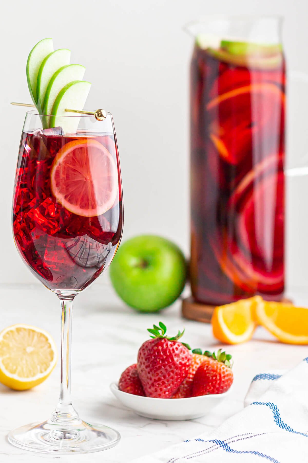 wine glass filled with non-alcoholic Sangria garnished with a green apple fan with a jug of Sangria in the background.
