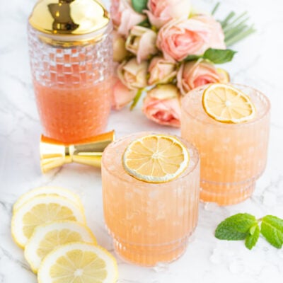 2 peach coloured drinks in short glasses topped with dehydrated lemon and a glass cocktail shaker with a gold lid in the background