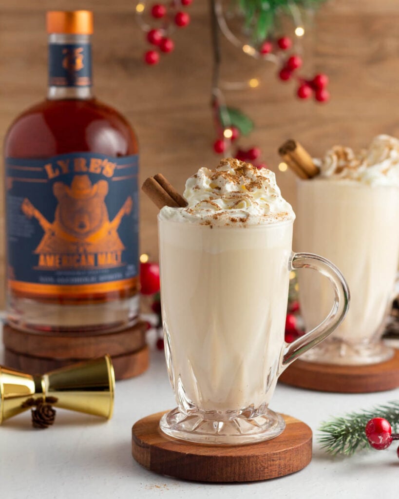 2 glasses of eggnog topped with whipped cream and a cinnamon stick, with a bottle of non alcoholic Lyre's American Malt in the background
