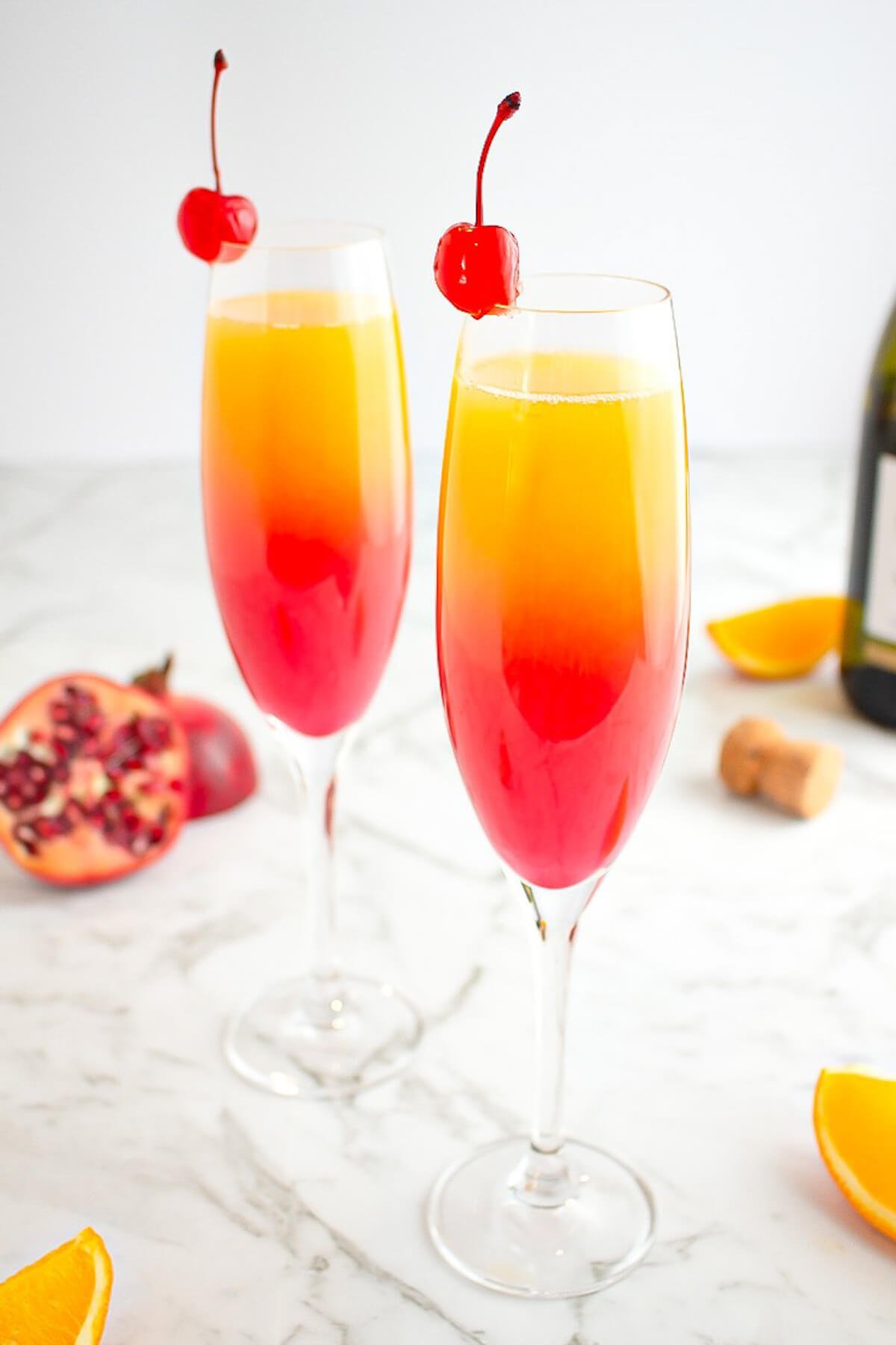 champagne glasses filled with a sunrise drink with red on the bottom progressing to orange then yellow, garnished with a maraschino cherry.