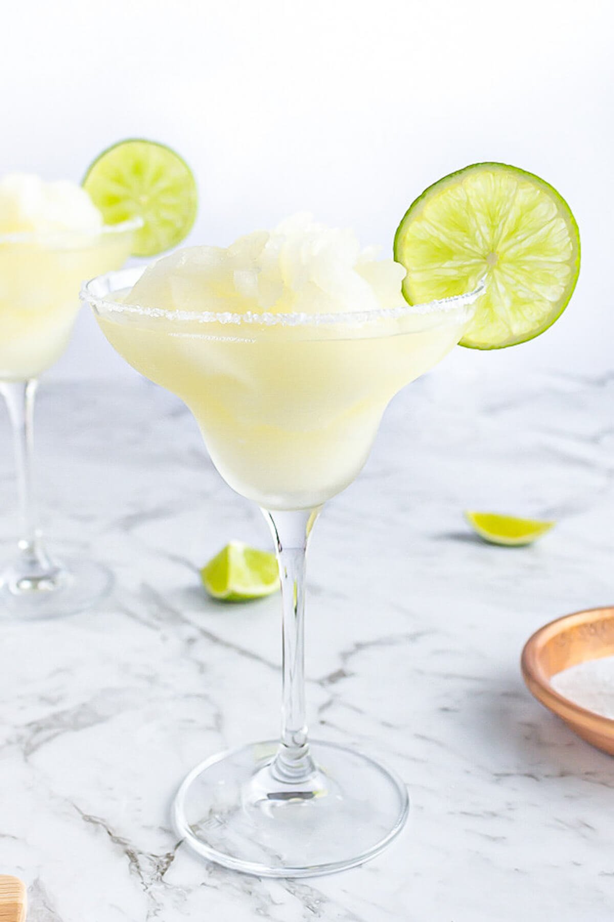 classic margarita glass filled with a frozen yellow slushie drink garnished with a lime wheel on a grey and white marble background.