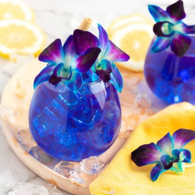 2 blue drinks in round glasses sitting on a light wooden board with a yellow cloth, garnished with purple and blue orchids
