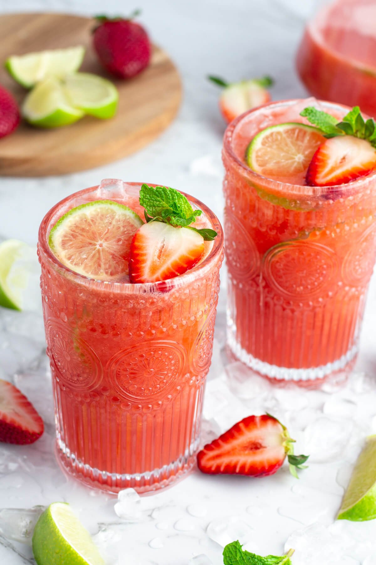 2 tall glasses of pink strawberry agua fresca garnished with a lime wheel, strawberry and sprig of mint on a white background.