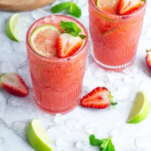 2 tall glasses of pink strawberry agua fresca garnished with a lime wheel, strawberry and sprig of mint on a white background