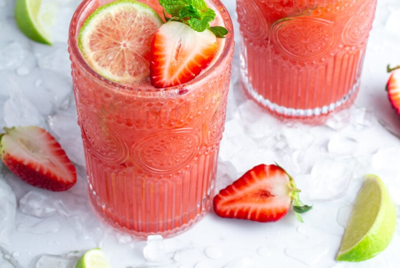 2 tall glasses of pink strawberry agua fresca garnished with a lime wheel, strawberry and sprig of mint on a white background